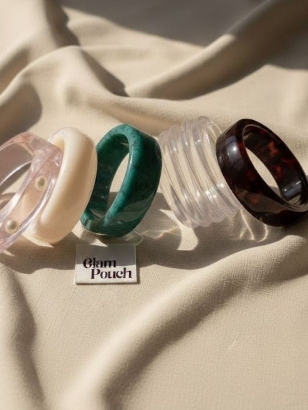 Five individual resin bangles laid flat on cream fabric next to a Glam Pouch branded card