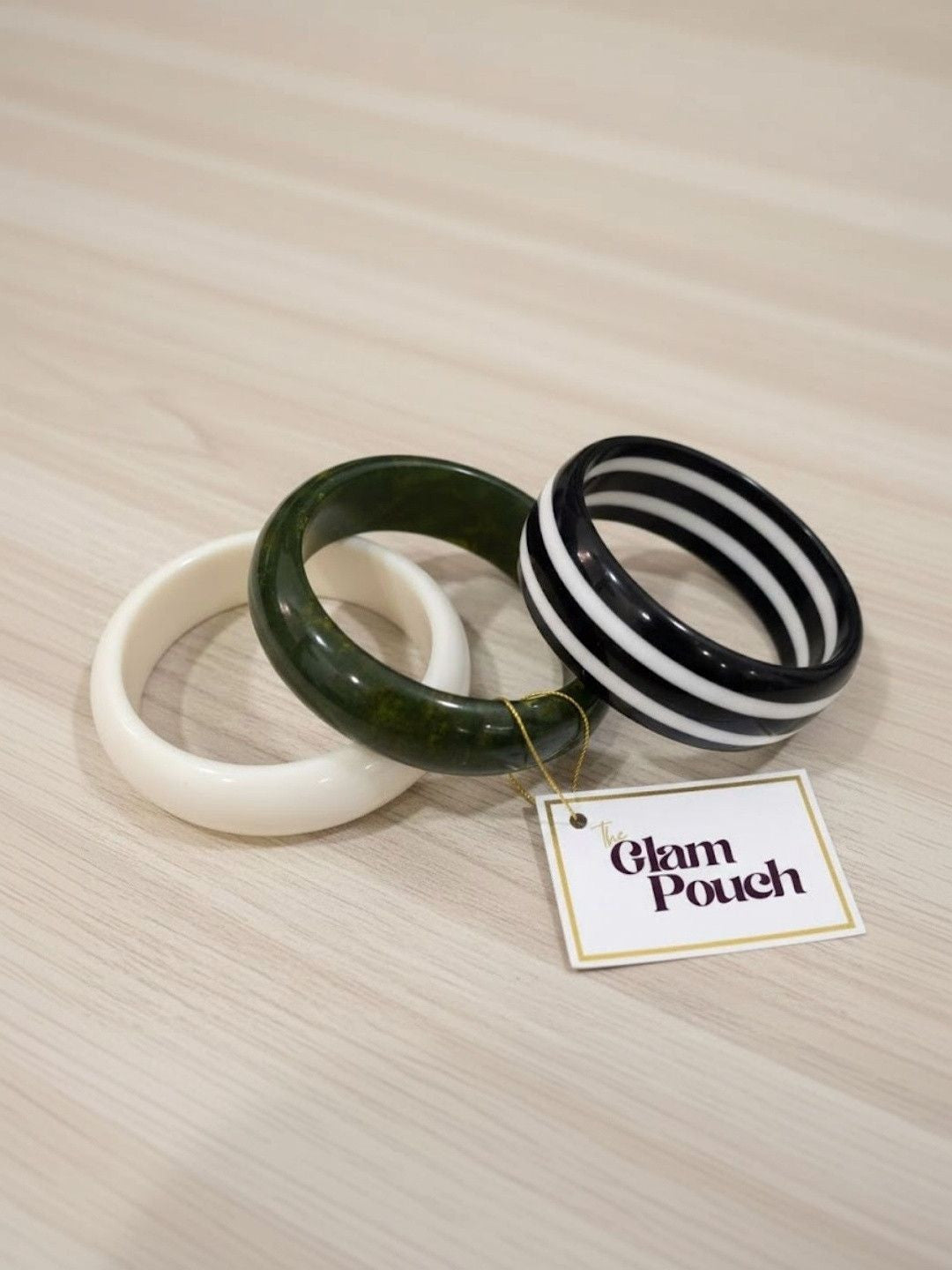 Three resin bangles laid flat on a wooden surface beside a Glam Pouch brand tag