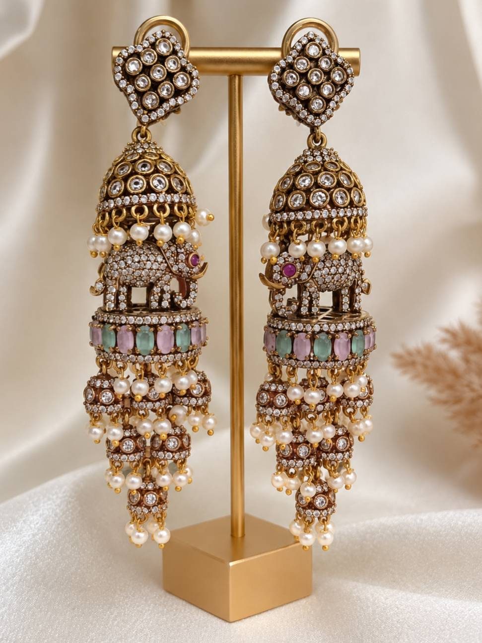 Pair of long oxidised gold elephant jhumka earrings displayed on a gold jewellery stand showing front view with pearl drops and pastel stones