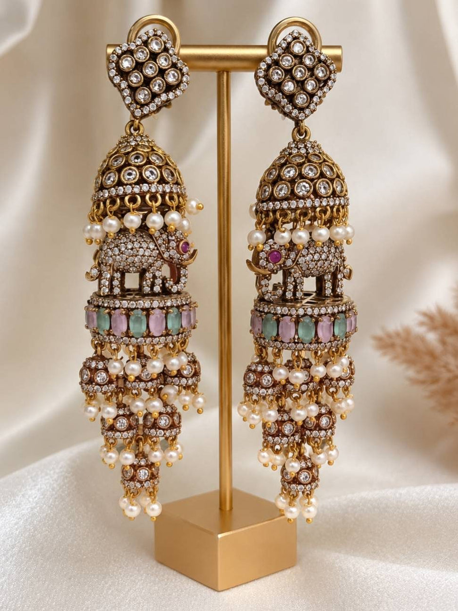 Pair of long oxidised gold elephant jhumka earrings displayed on a gold jewellery stand showing front view with pearl drops and pastel stones