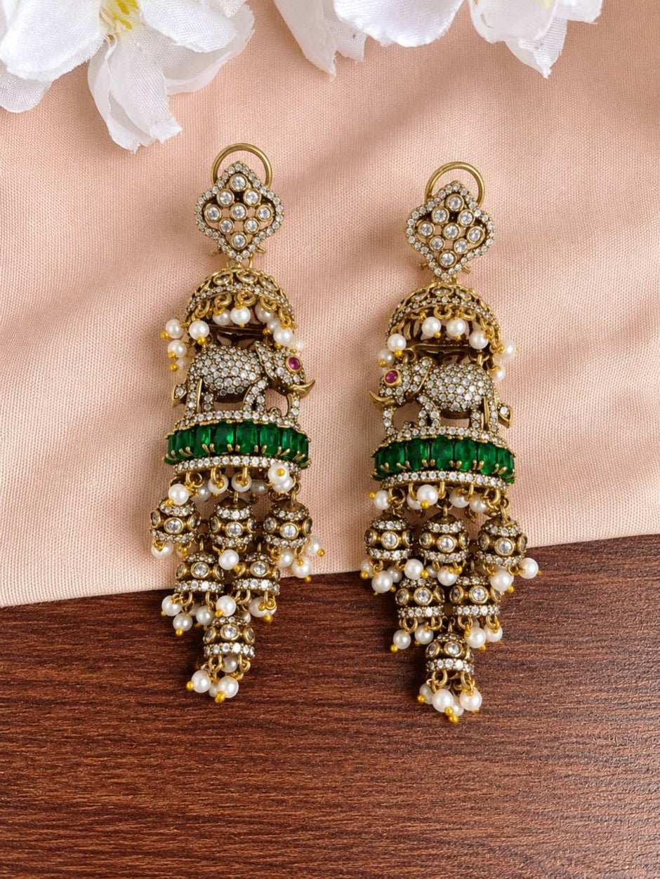 Front view of both antique gold elephant jhumka earrings laid flat on blush fabric with white flowers, showing full tiered design