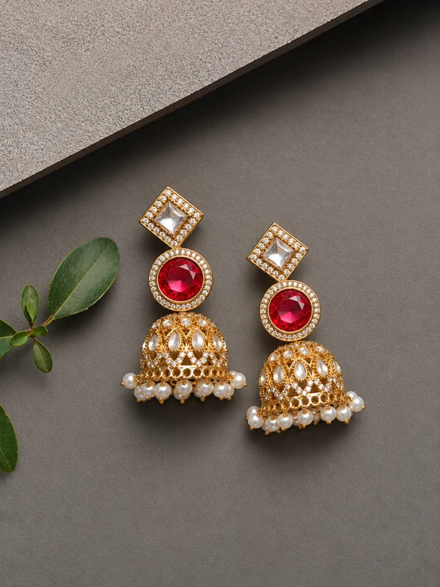 Pair of gold-plated kundan jhumka earrings with ruby centre stone and pearl drops laid flat on grey surface