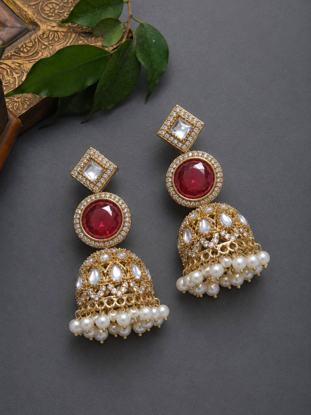 Pair of gold-plated kundan ruby jhumka earrings with pearl fringe displayed on grey surface