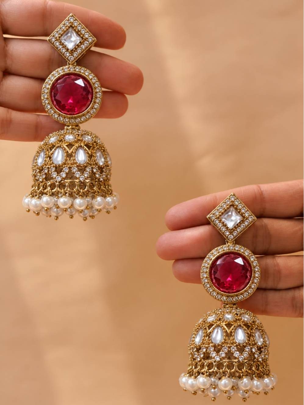 Two hands holding matching gold-plated kundan ruby jhumka earrings showing full design detail