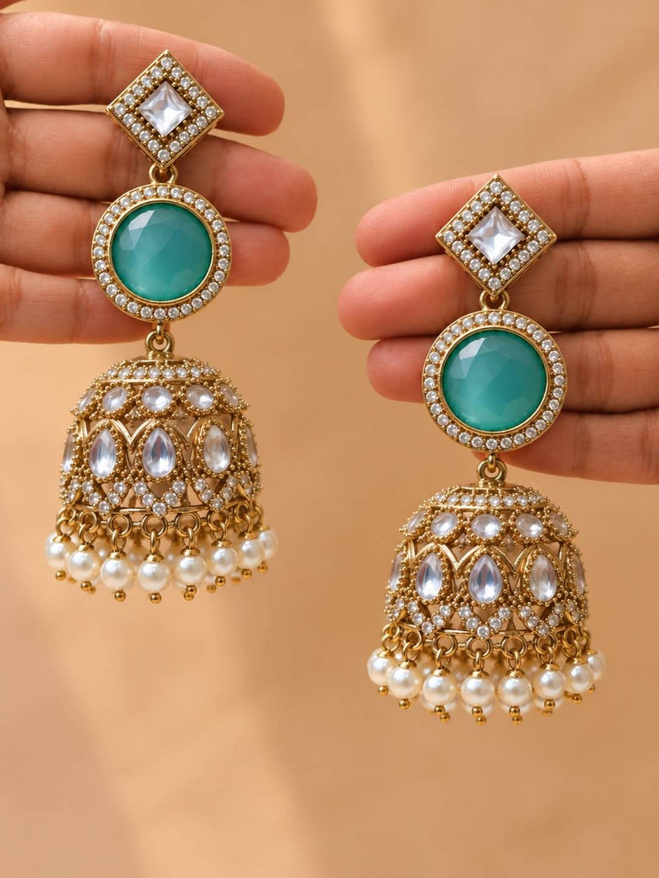 Two hands each holding one gold-plated kundan jhumka earring displaying the three-tier design with turquoise stone and pearls