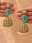Two hands each holding one gold-plated kundan jhumka earring displaying the three-tier design with turquoise stone and pearls