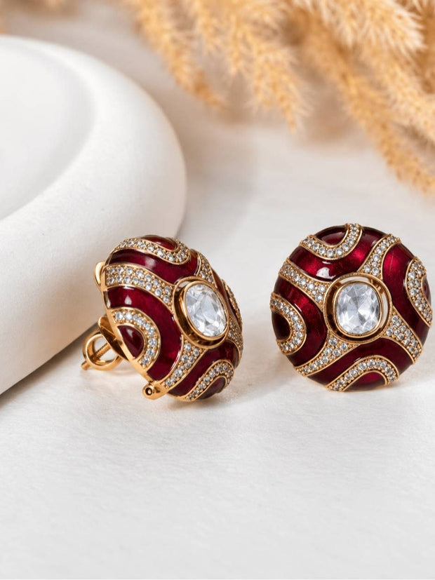 Pair of red enamel and gold-tone kundan dome stud earrings displayed on a white surface with dried pampas grass in the background