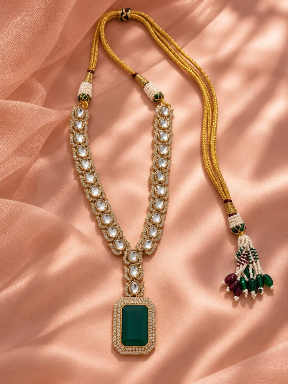 Long mala-style kundan necklace with emerald green rectangular pendant on golden dori cord with pearl and bead tassel laid on peach fabric