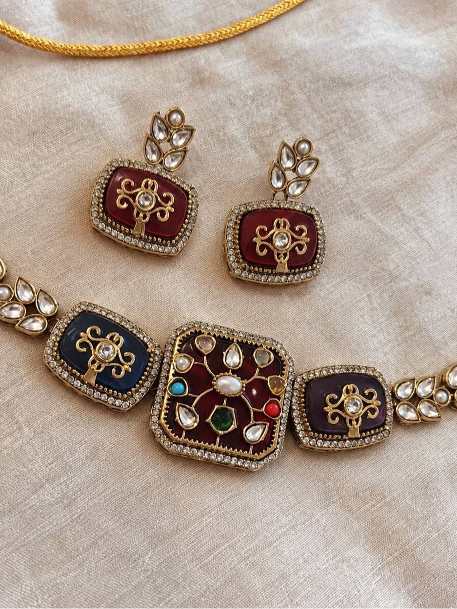 Close-up flat lay of kundan choker necklace and pair of maroon enamel earrings on cream fabric showing intricate stone and enamel detailing