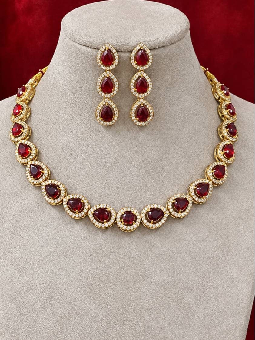 Gold-plated ruby CZ teardrop necklace and triple-drop earrings displayed on a grey jewellery bust against red velvet background