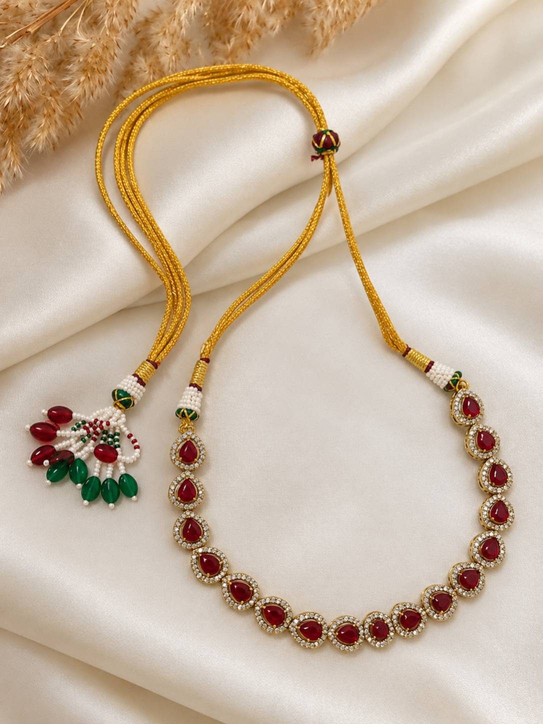 Long gold-plated ruby CZ teardrop necklace with golden silk thread adjustable cord and decorative beaded tassel, laid flat on ivory fabric