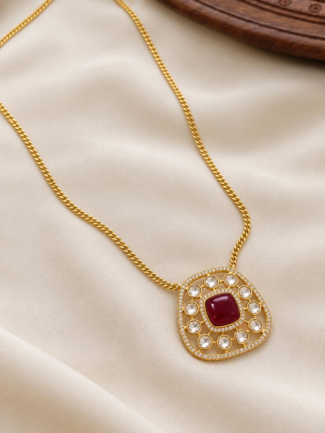 Close-up of gold-plated cushion pendant with red cabochon centre, kundan accents and pavé border on a curb chain laid on cream silk fabric