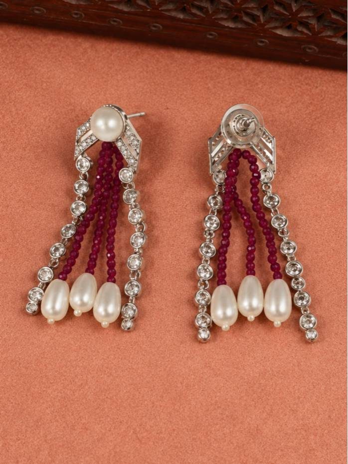 Close-up of silver-tone crystal and ruby bead drop earrings with teardrop pearl ends on a terracotta background