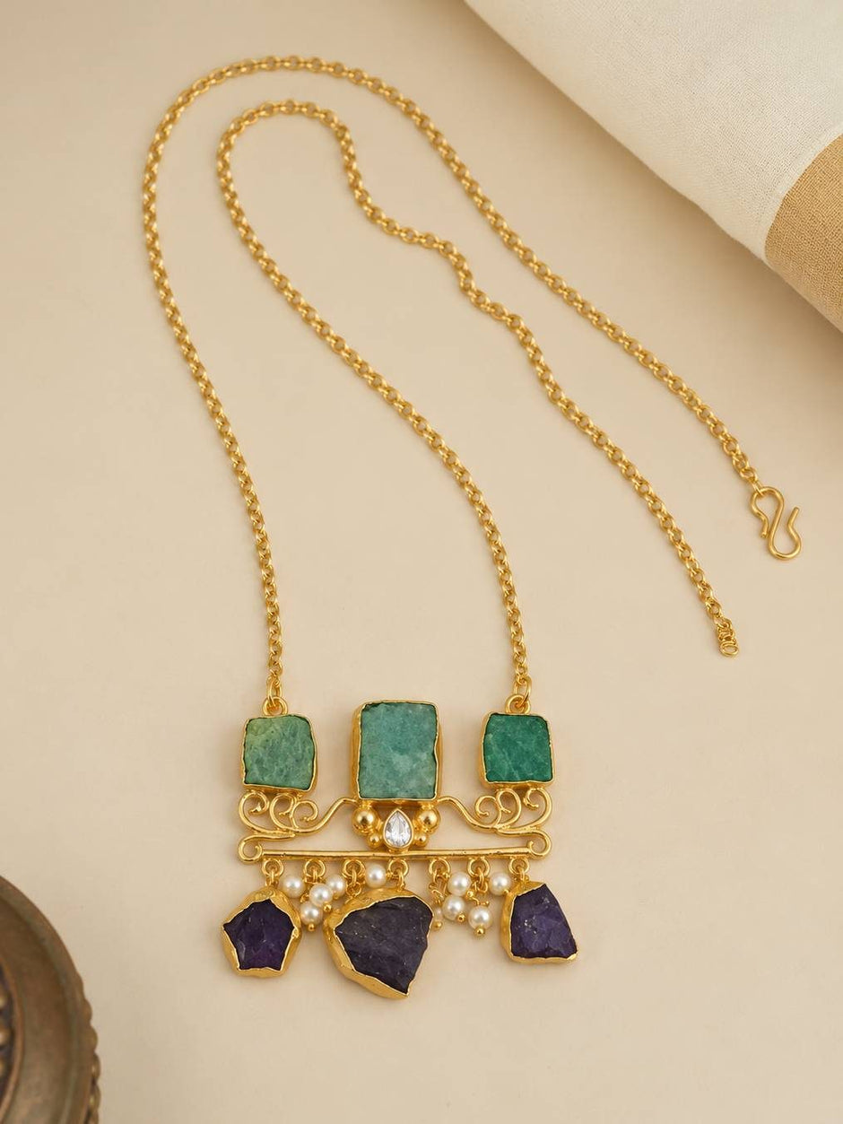 Flat lay of gold-plated long chain statement necklace with three green stone squares and hanging purple amethyst chunks and seed pearls
