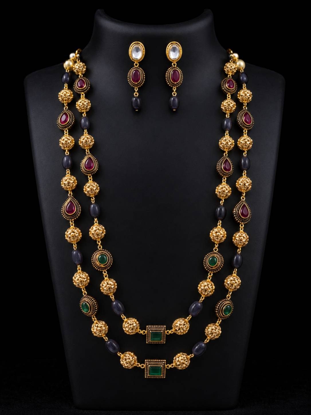 Gold-plated double-strand beaded necklace set with ruby and emerald colour stones displayed on a black mannequin bust with matching drop earrings