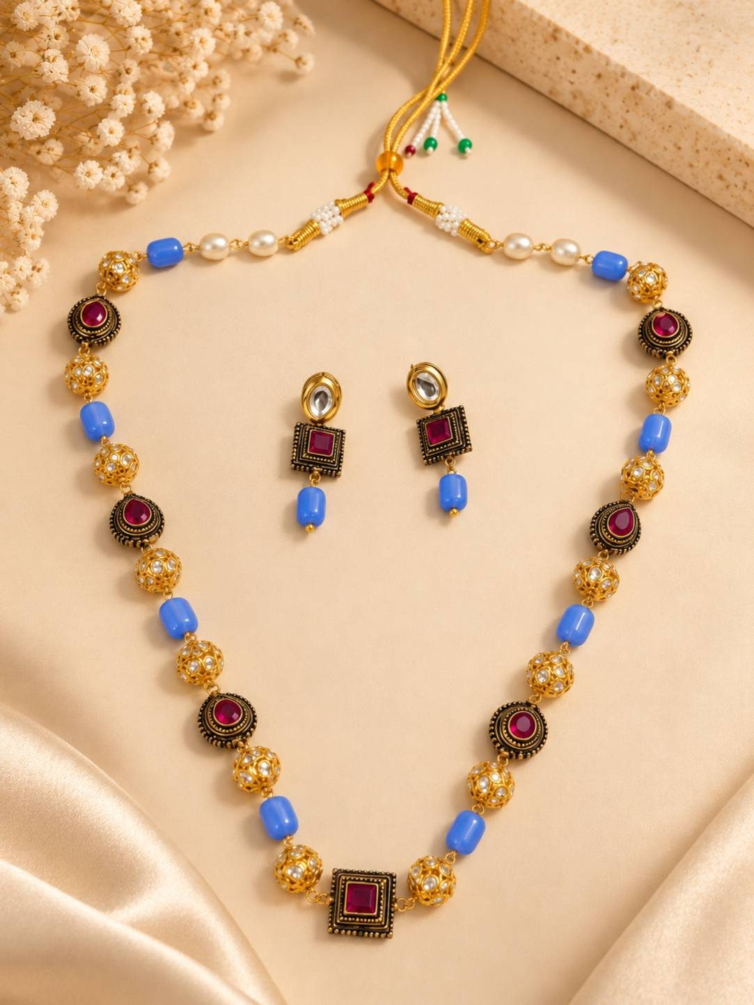 Gold-plated kundan beaded necklace set with blue barrel beads and matching drop earrings laid flat on cream fabric with dried flowers