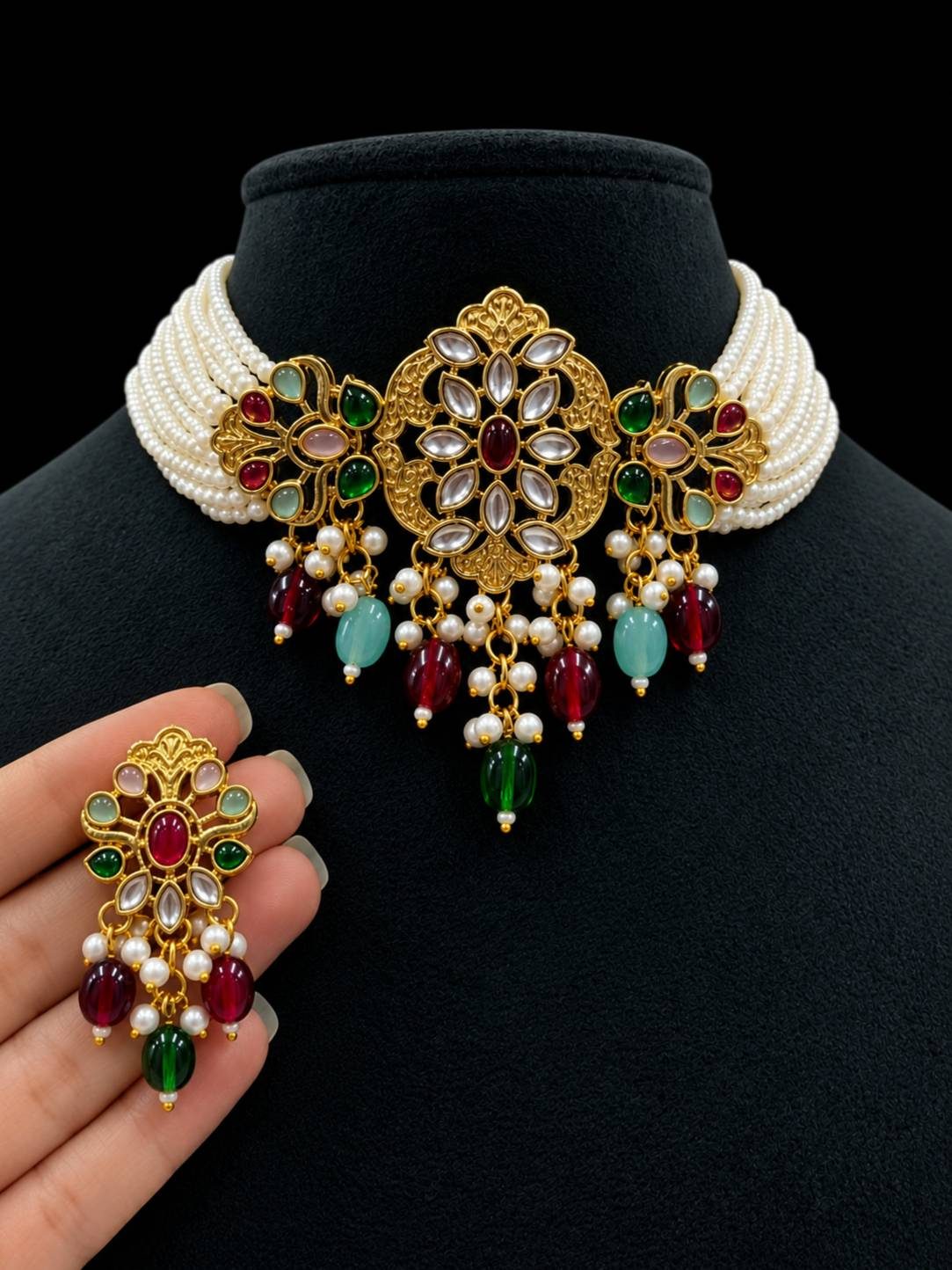 Kundan pearl choker necklace displayed on a black bust with matching earring held in hand, showing red, green and aqua stones
