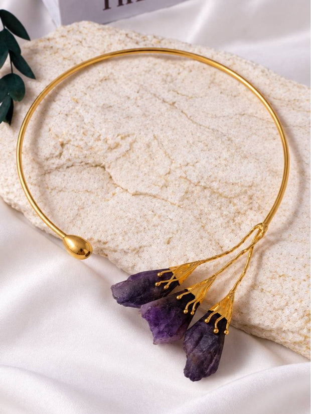 Gold-plated open torque choker with raw purple amethyst stone drops laid flat on a textured white stone surface