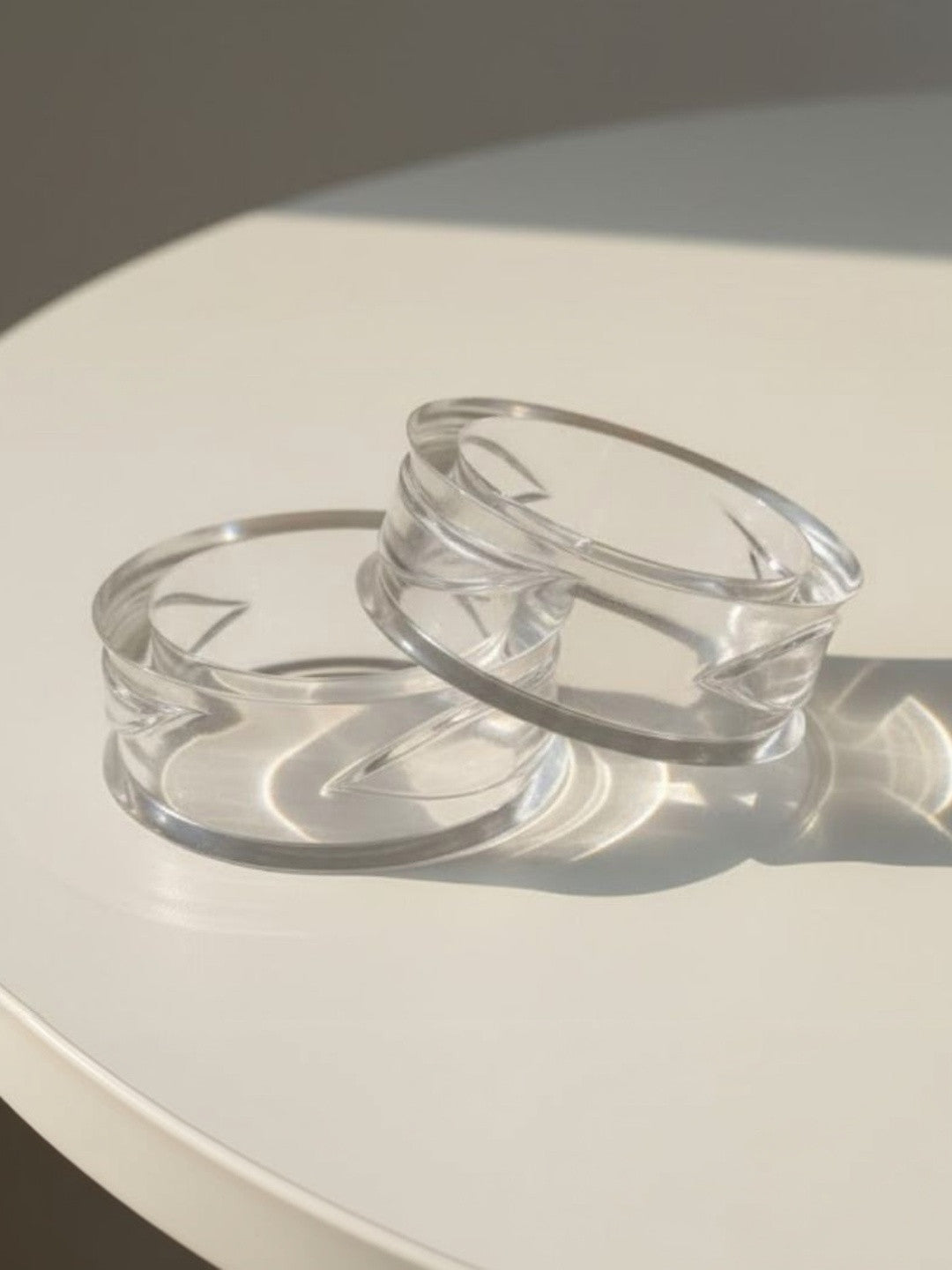 Two clear geometric acrylic bangles resting on a white surface in natural sunlight