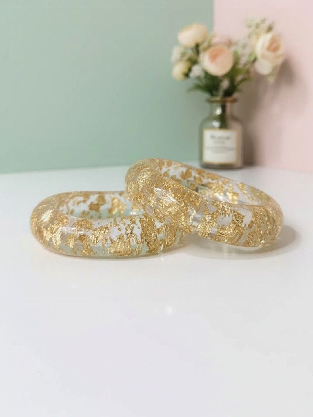 Two clear resin bangles with scattered gold foil flakes resting on a white surface against a mint and pink background