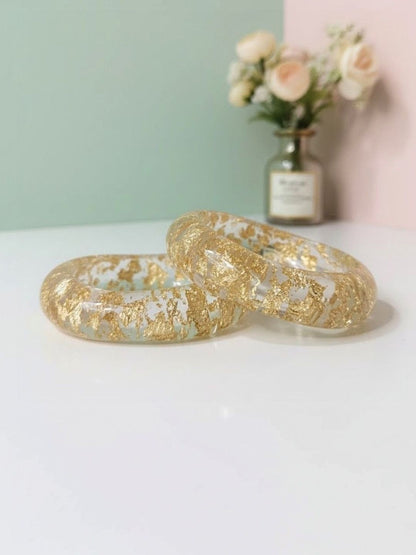 Two clear resin bangles with scattered gold foil flakes resting on a white surface against a mint and pink background