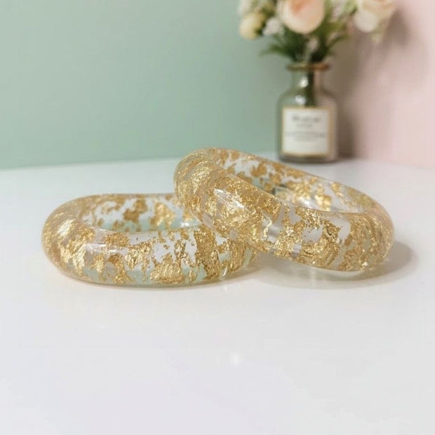 Two clear resin bangles with scattered gold foil flakes resting on a white surface against a mint and pink background