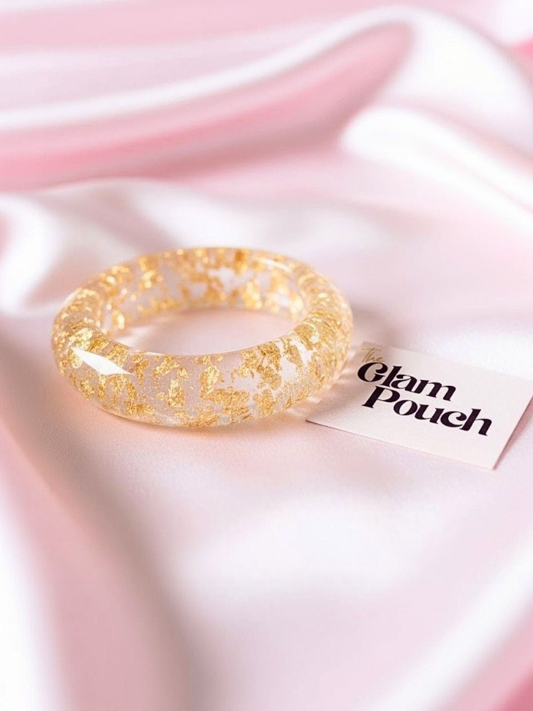 Single clear resin bangle with gold foil inclusions placed on pink satin fabric beside a Glam Pouch brand card
