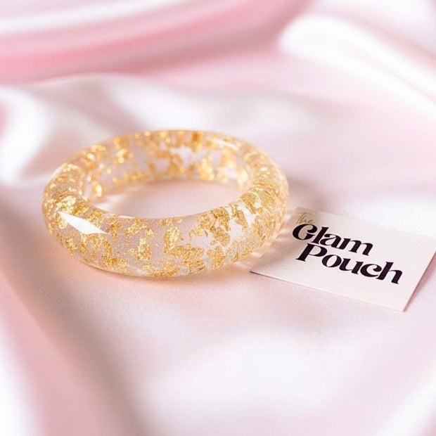 Single clear resin bangle with gold foil inclusions placed on pink satin fabric beside a Glam Pouch brand card