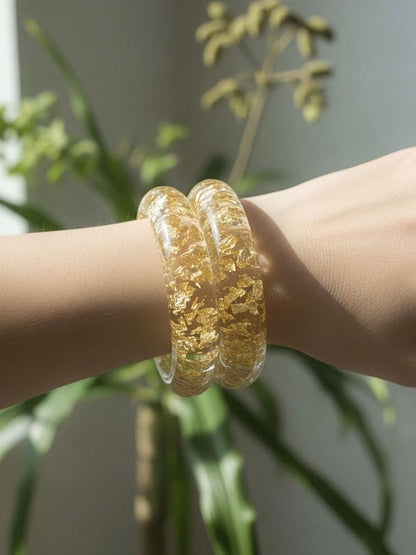 Two gold foil clear resin bangles stacked and worn on a woman's wrist with green foliage in the background