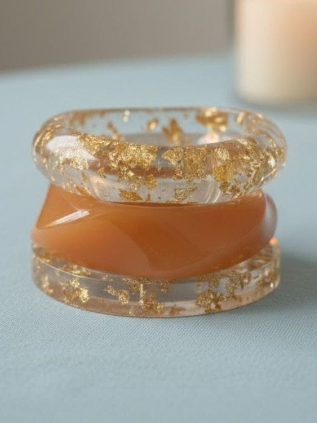 Three stacked bangles on light blue surface — two clear resin with gold foil flakes and one peach opaque resin bangle
