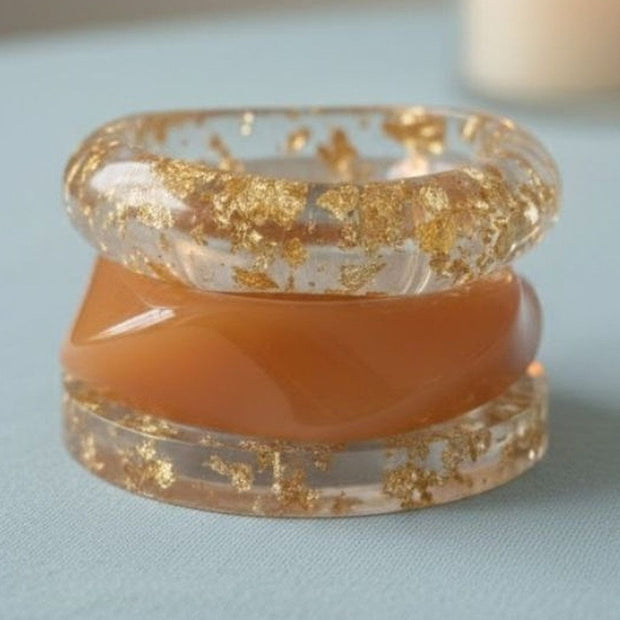 Three stacked bangles on light blue surface — two clear resin with gold foil flakes and one peach opaque resin bangle