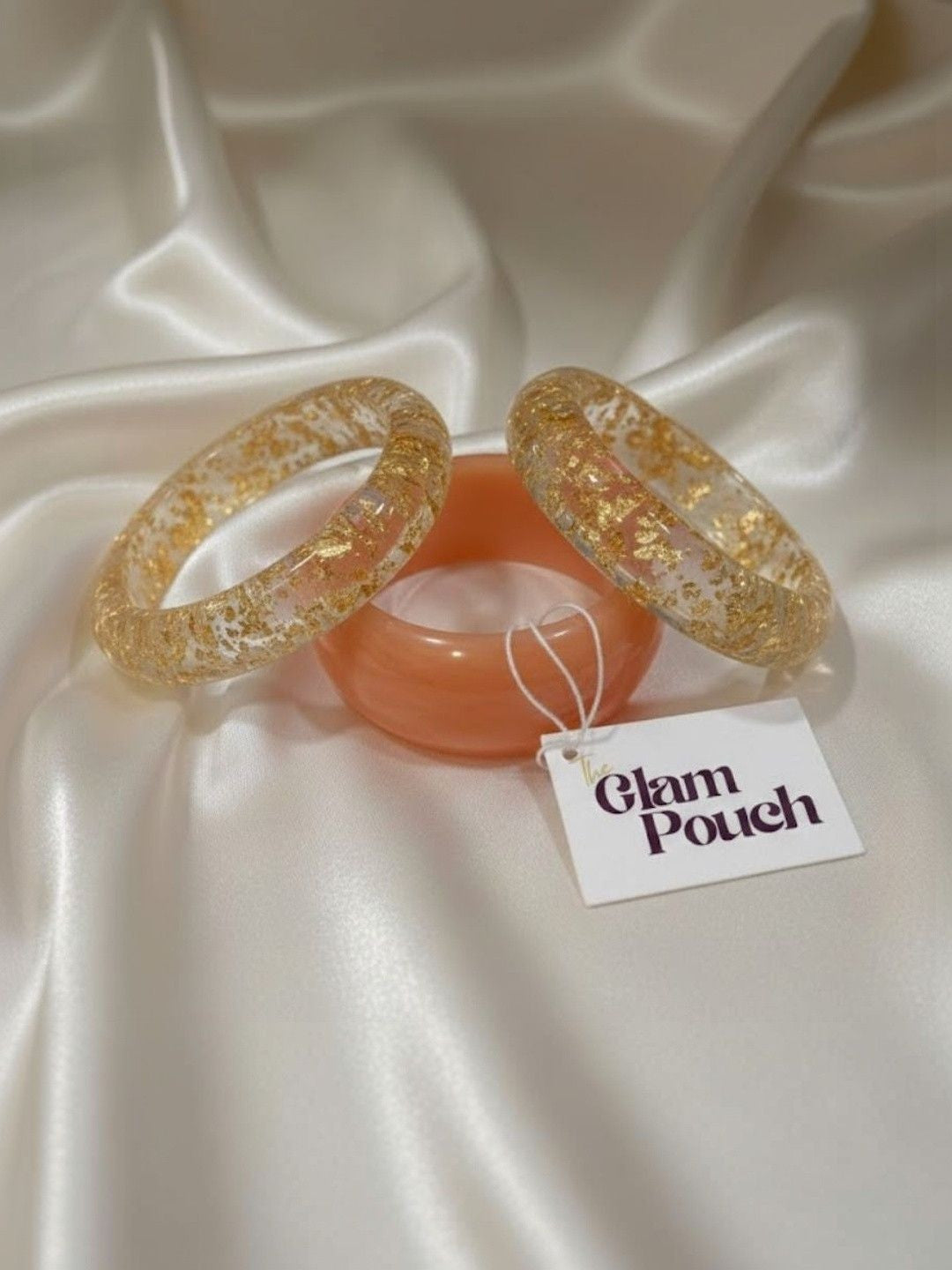 Flatlay of three bangles on white satin with Glam Pouch tag — two clear gold foil resin and one peach resin bangle