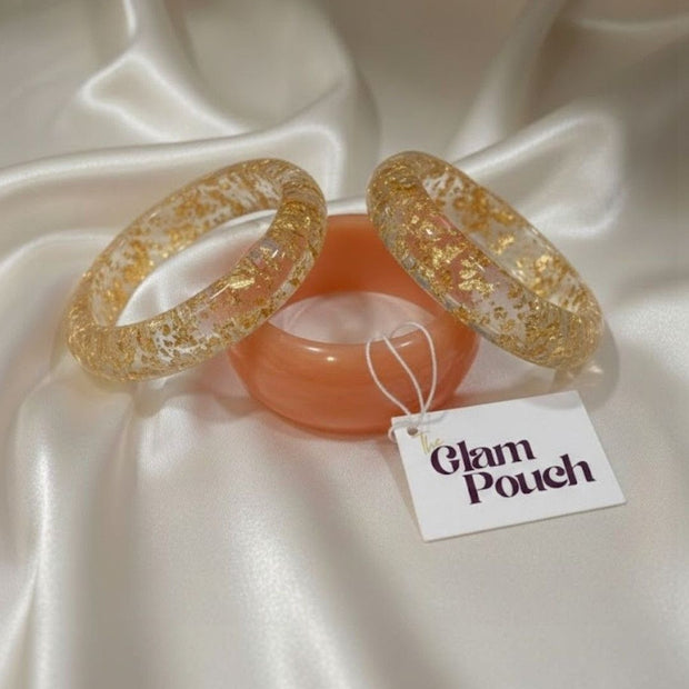 Flatlay of three bangles on white satin with Glam Pouch tag — two clear gold foil resin and one peach resin bangle