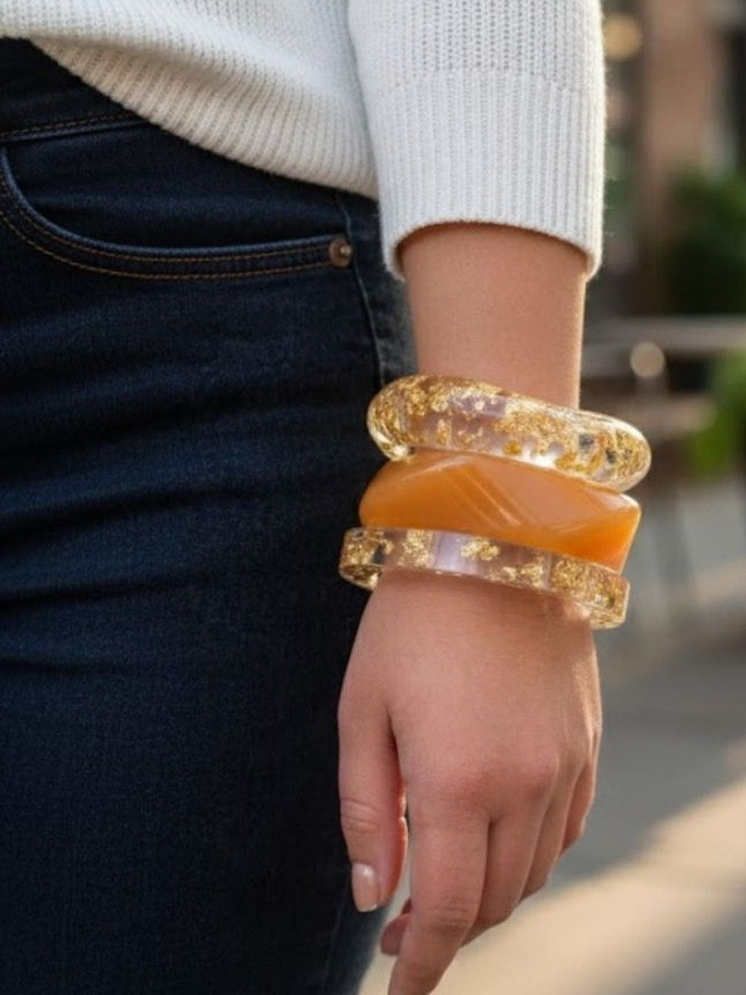 Woman wearing three stacked bangles on wrist — clear gold foil resin bangles layered with a peach opaque bangle, styled with white sweater and dark jeans