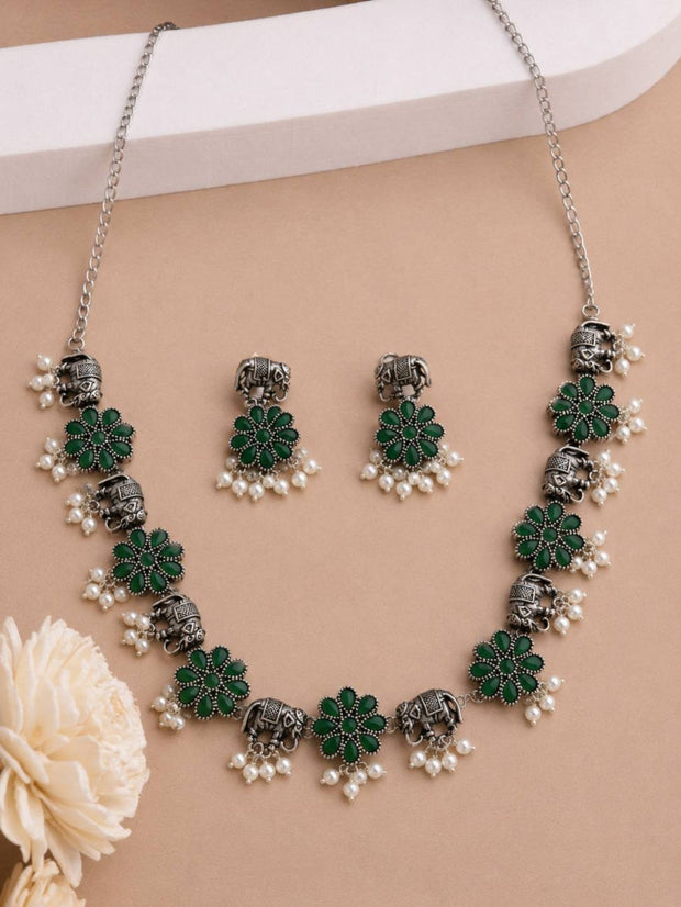 Flat lay of oxidised silver floral necklace and elephant earring set with green stones and faux pearls on a beige background with white flower