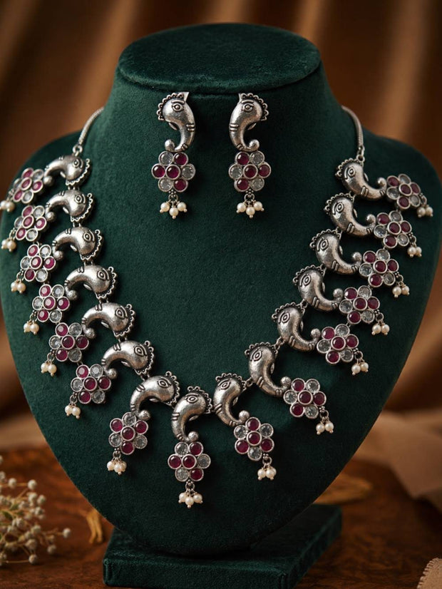 Oxidised silver peacock necklace set with ruby and clear stone floral drops displayed on a dark green velvet jewellery bust