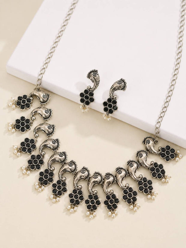 Peacock motif oxidised silver choker necklace and stud earrings laid flat on a cream surface beside a white block