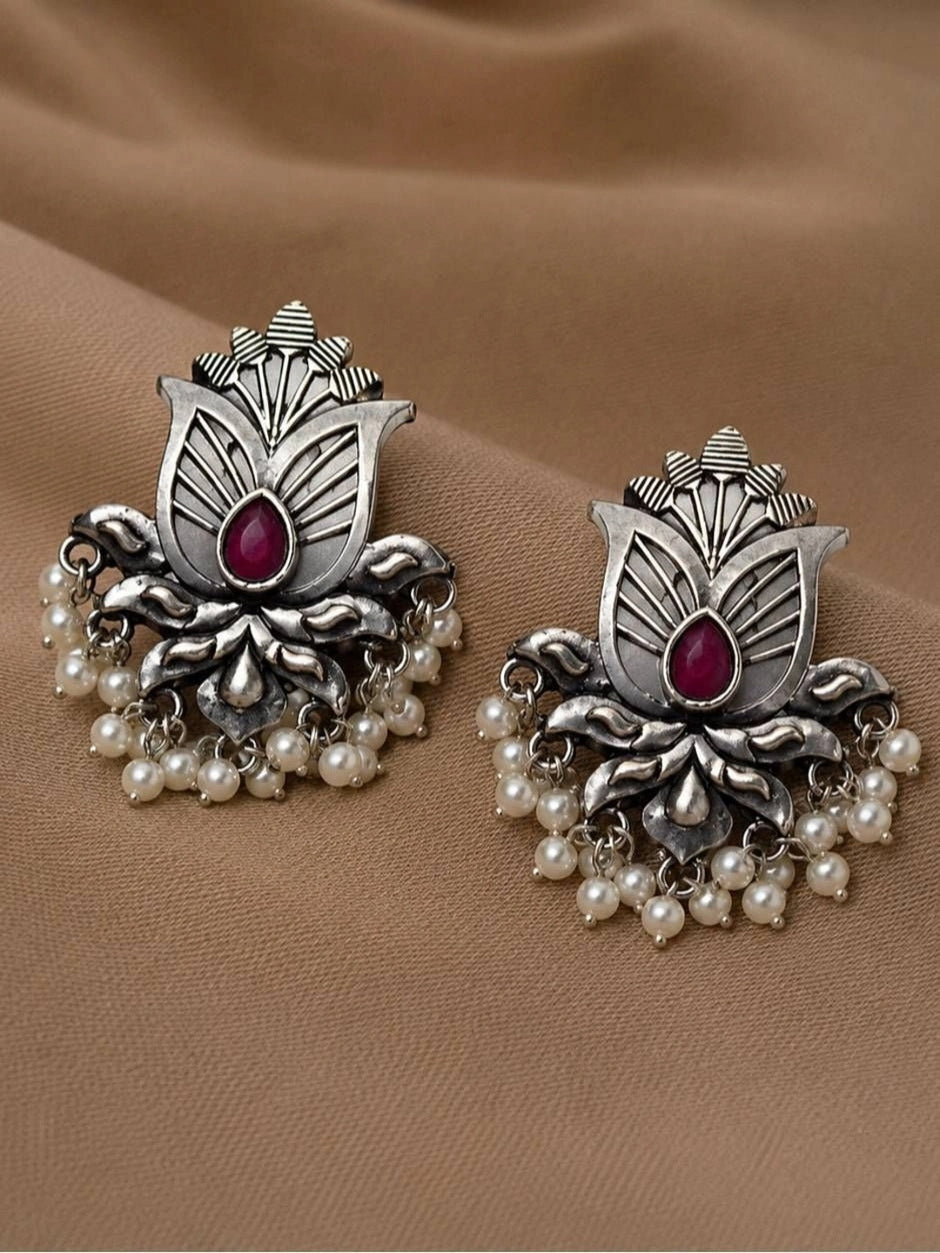 Pair of oxidised silver lotus stud earrings with ruby-red teardrop stone and white pearl drops, laid flat on beige fabric