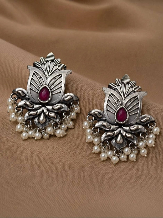 Pair of oxidised silver lotus stud earrings with ruby-red teardrop stone and white pearl drops, laid flat on beige fabric