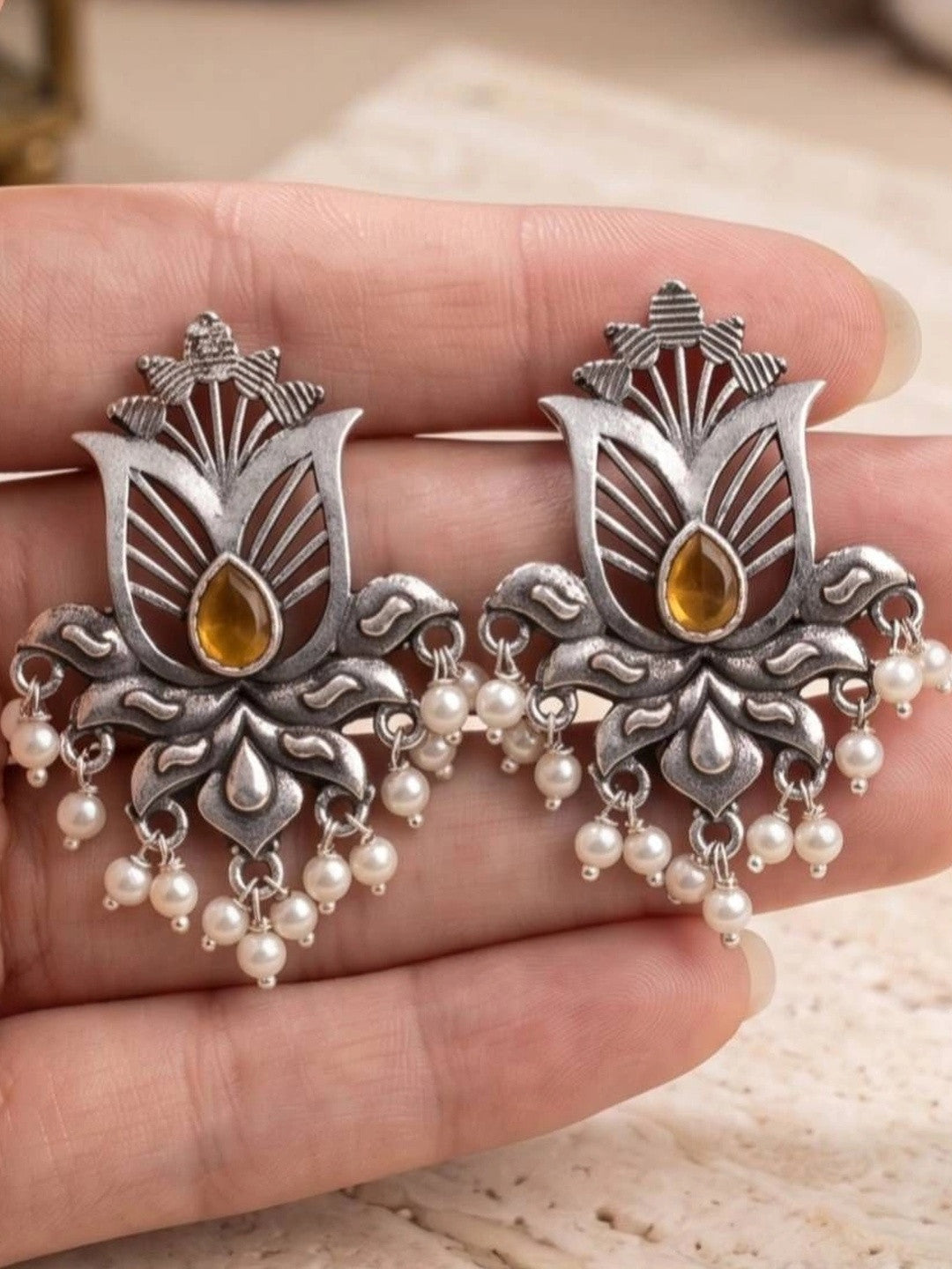 Close-up of two oxidised silver lotus earrings with amber stone centres and pearl drops held in palm