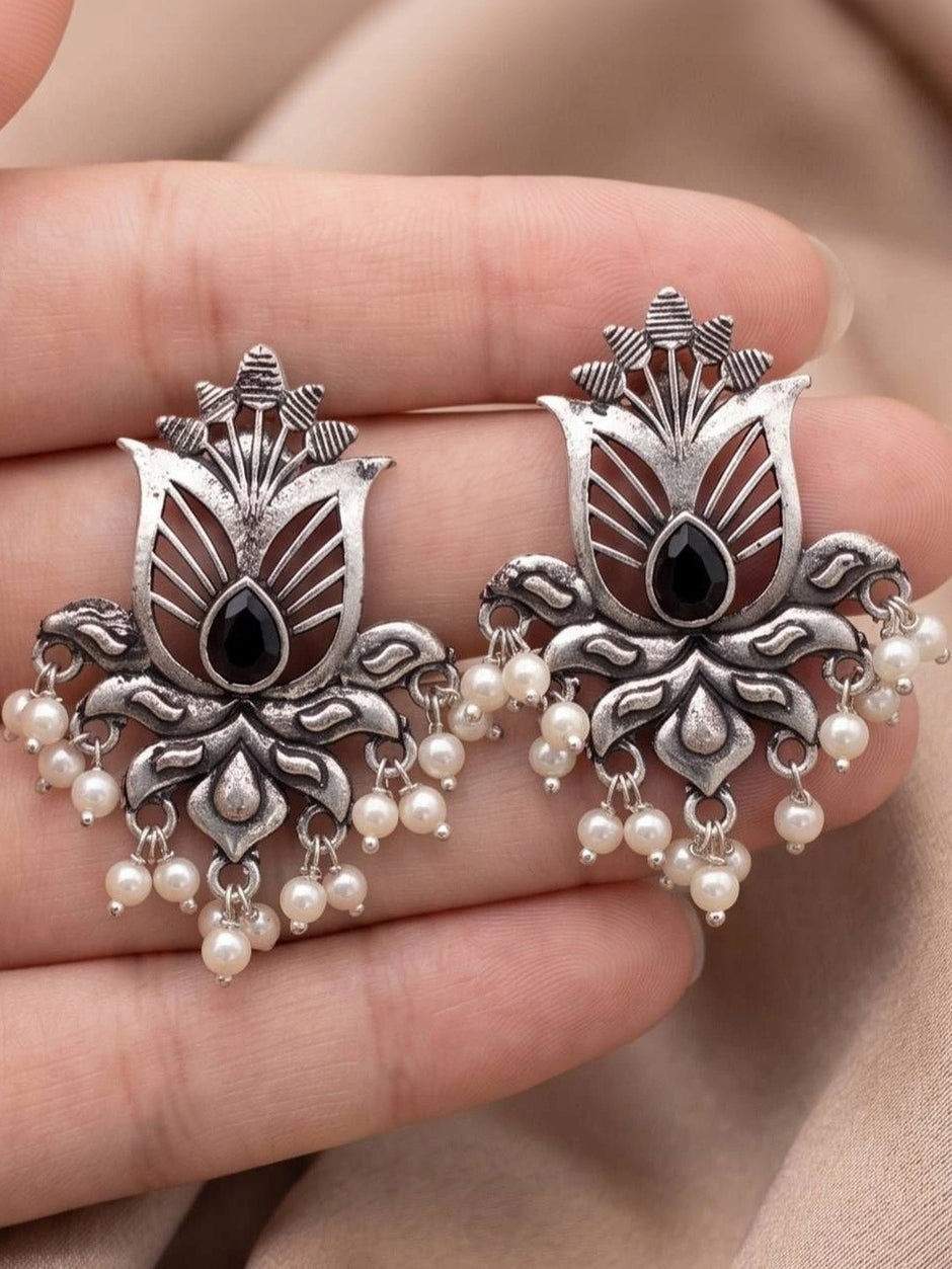 Two oxidised silver lotus stud earrings held in a palm showing open-cut design, black centre stone and pearl fringe