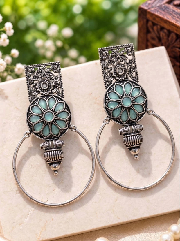 Pair of oxidised silver floral hoop earrings with aqua glass lotus motif on a cream surface
