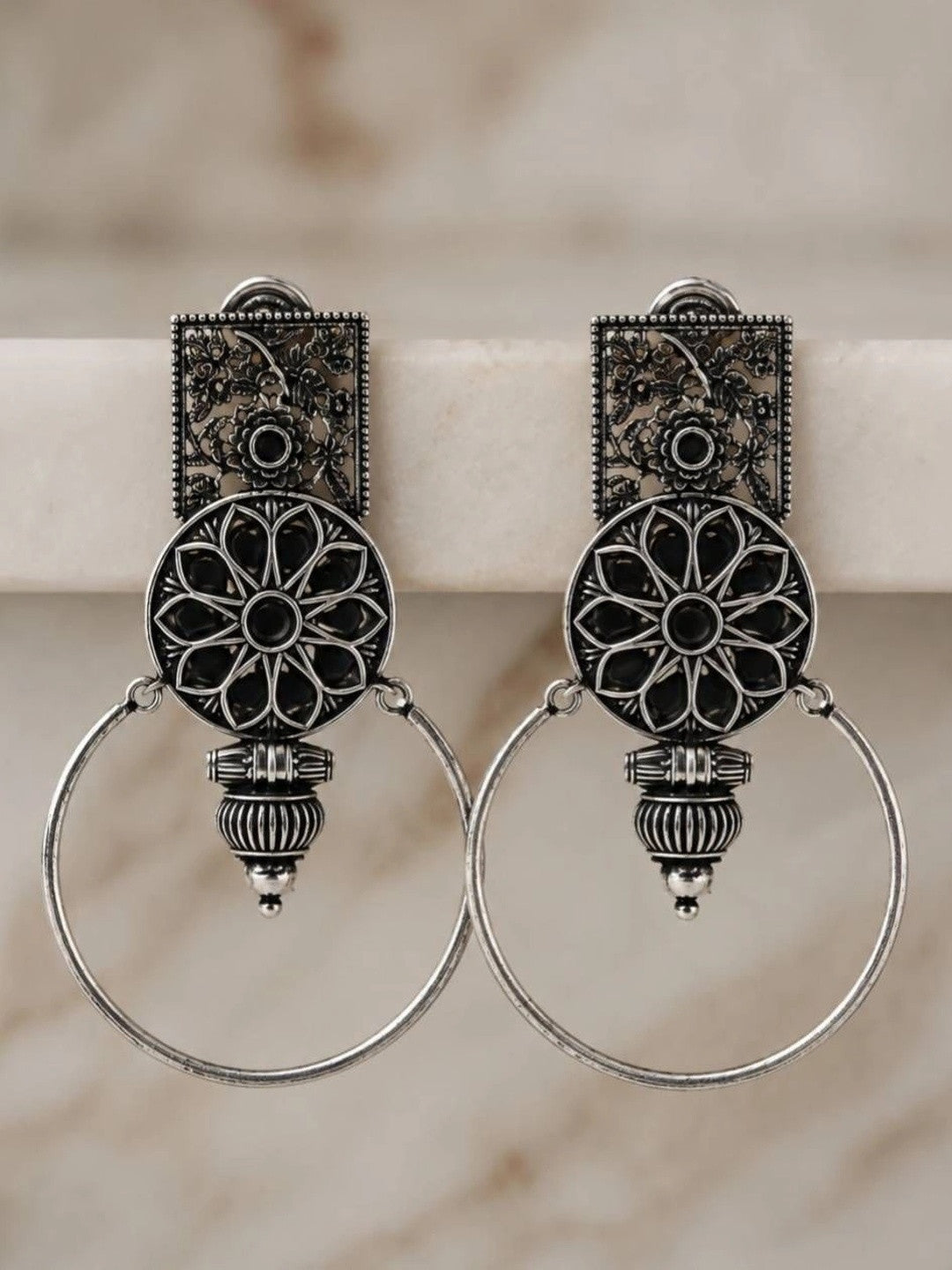 Pair of oxidised silver floral hoop earrings with black resin mandala disc displayed upright against a marble background