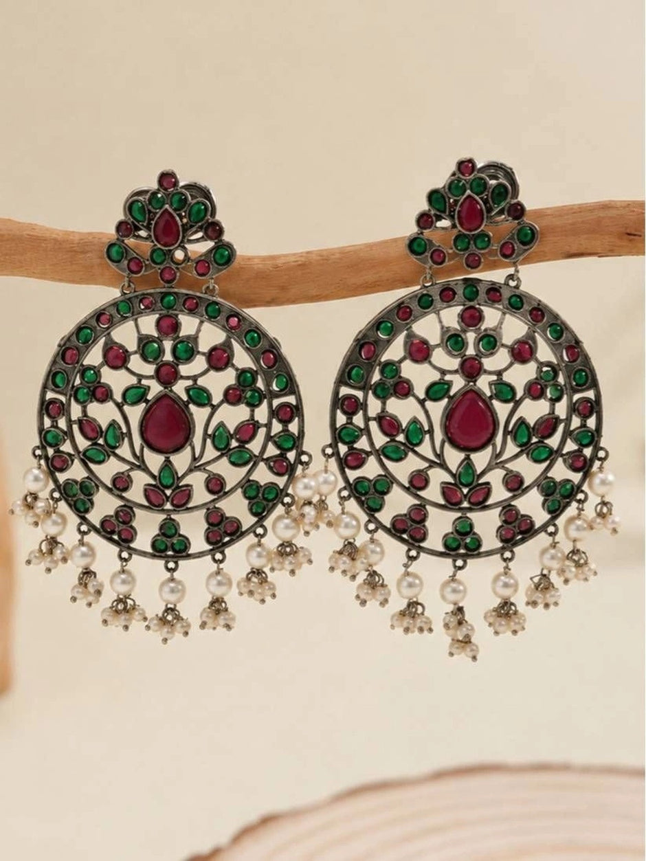 Pair of oxidised silver chandbali earrings with ruby-red and green stones and pearl drops, displayed on a wooden branch against a beige background