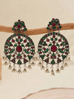 Pair of oxidised silver chandbali earrings with ruby-red and green stones and pearl drops, displayed on a wooden branch against a beige background