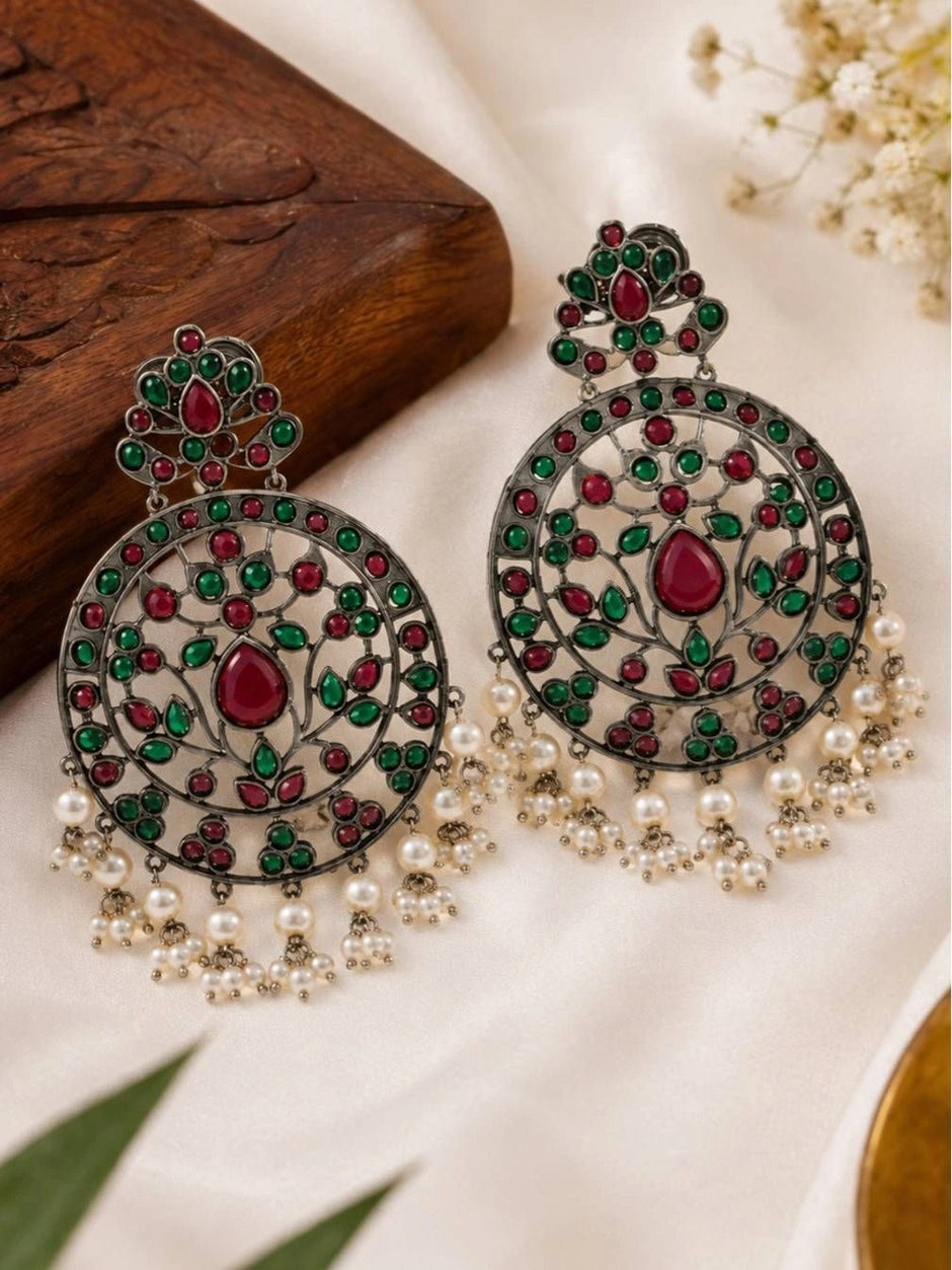 Two oxidised silver chandbali earrings laid flat on white fabric beside a wooden block and small white flowers