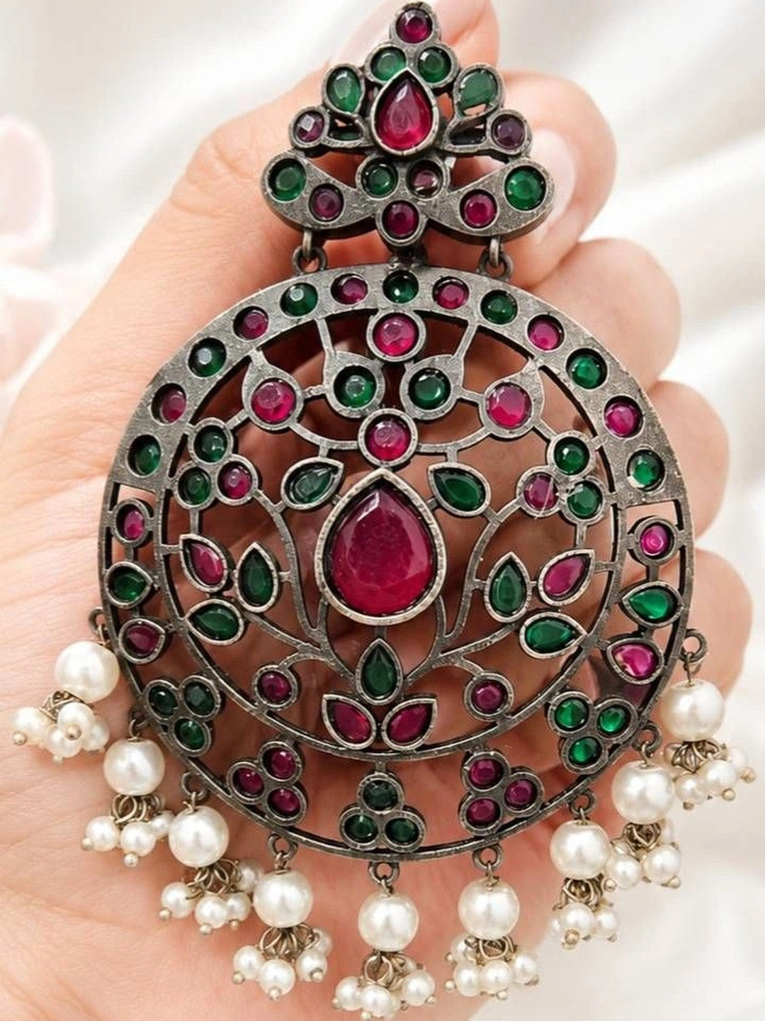 Single oxidised chandbali earring held in a hand, showing detailed ruby and green stone setting and cascading pearl drops