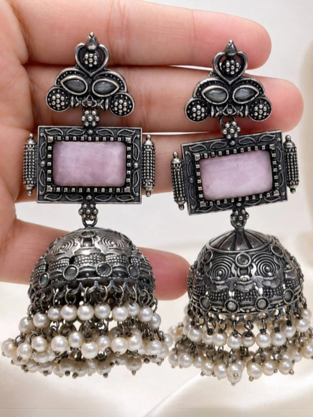 Close-up of oxidised silver jhumka earring pair held in a hand showing engraved dome and pink stone
