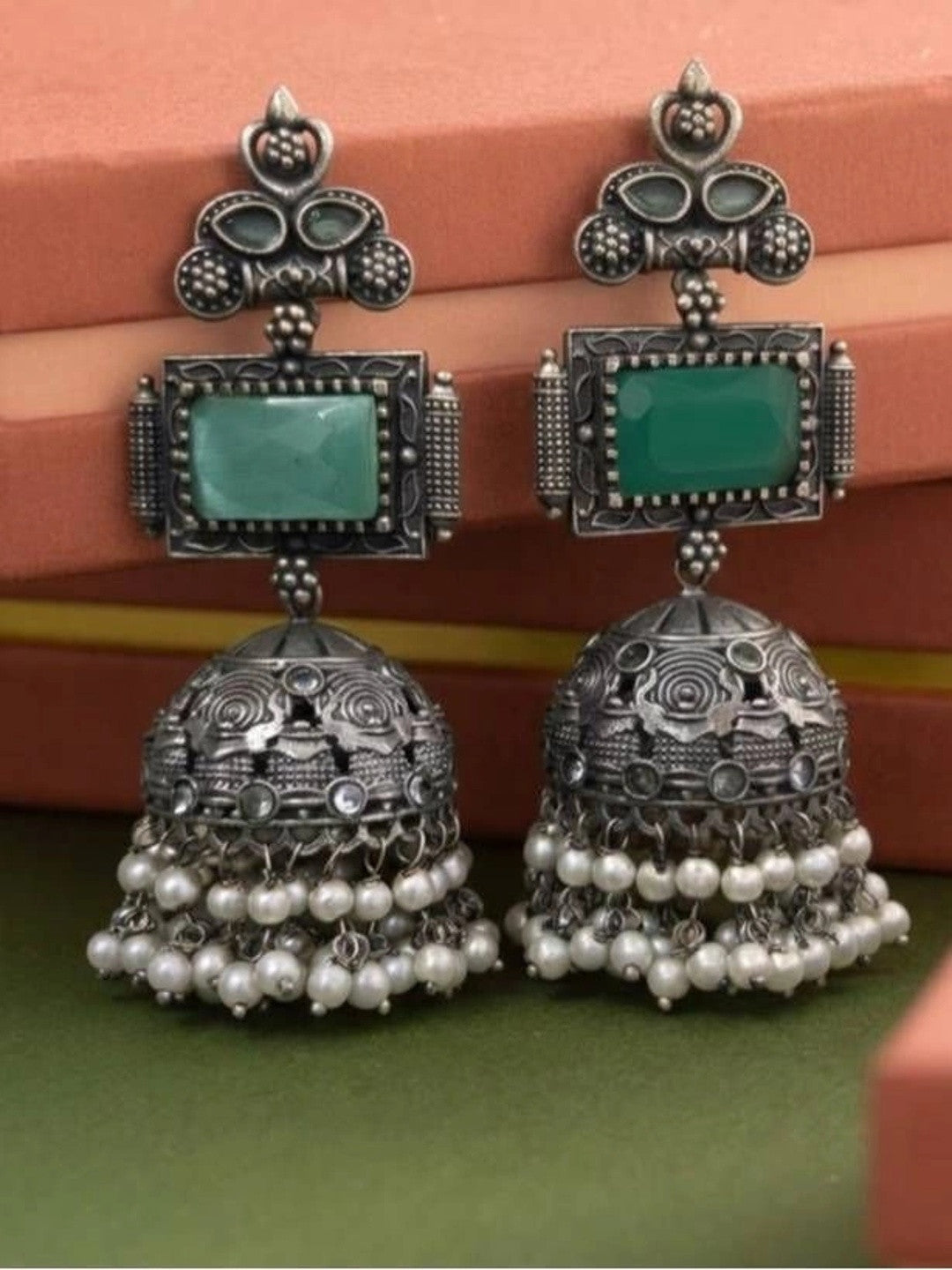 Pair of oxidised silver jhumka earrings with green rectangular stone and white pearl bead fringe displayed on green surface