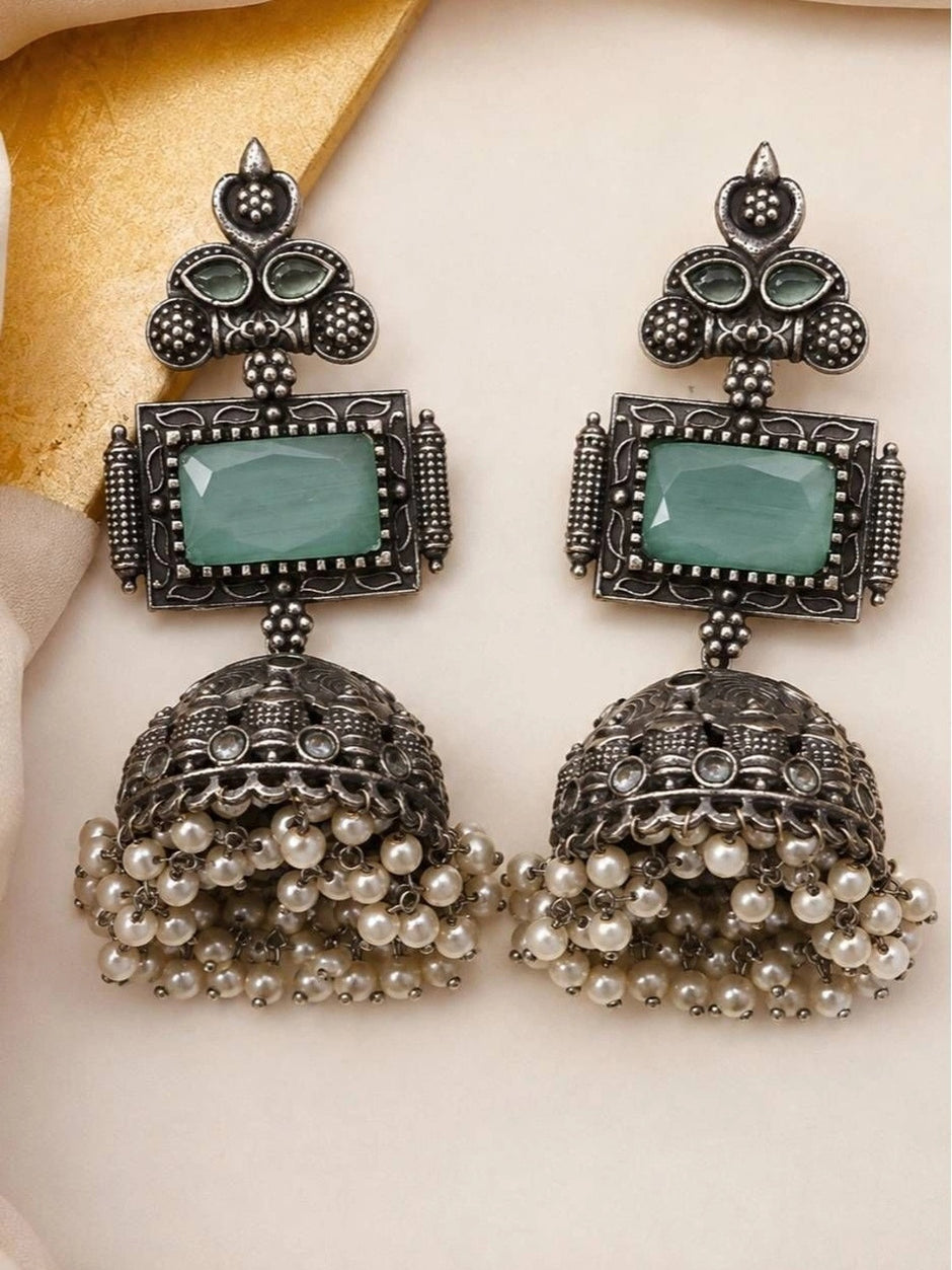 Front-facing pair of oxidised silver jhumka earrings with faceted green stone and pearl dangles on cream background